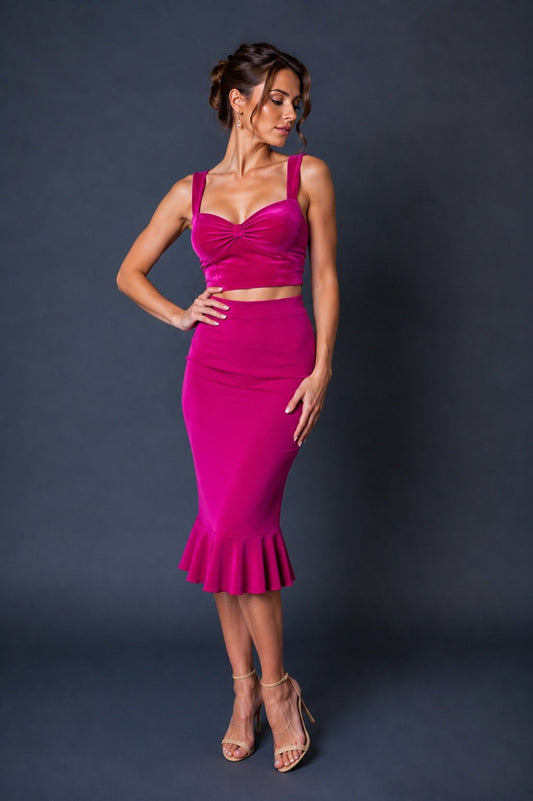 A woman modeling a bright fuchsia pink two-piece set consisting of a ruched crop top and a high-waisted flared midi skirt, paired with nude high-heeled sandals against a dark gray studio backdrop.