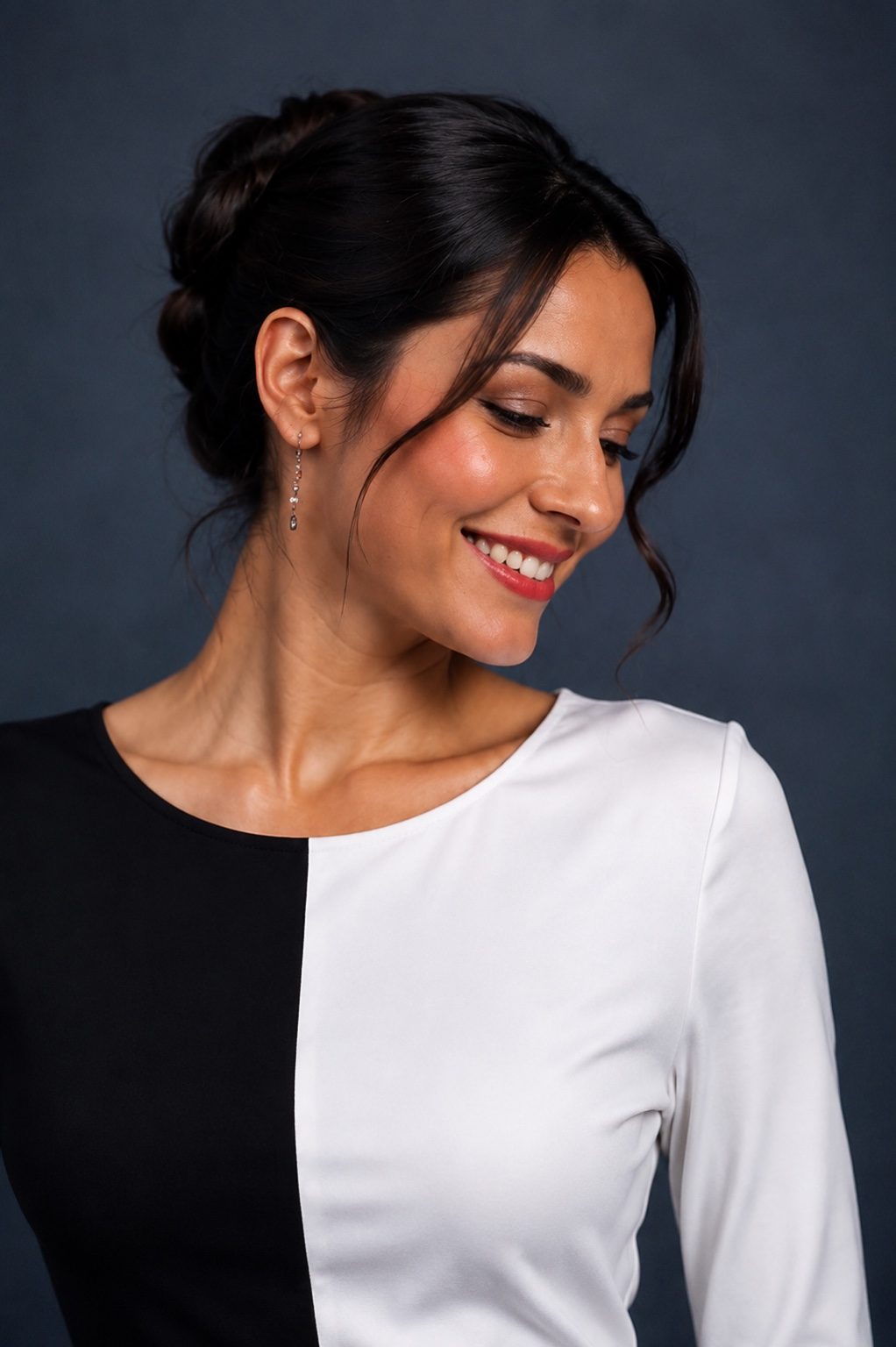 A woman with dark hair in a half-up bun wearing a long-sleeve top with a vertical split design in black and white, posing against a dark gray studio background.