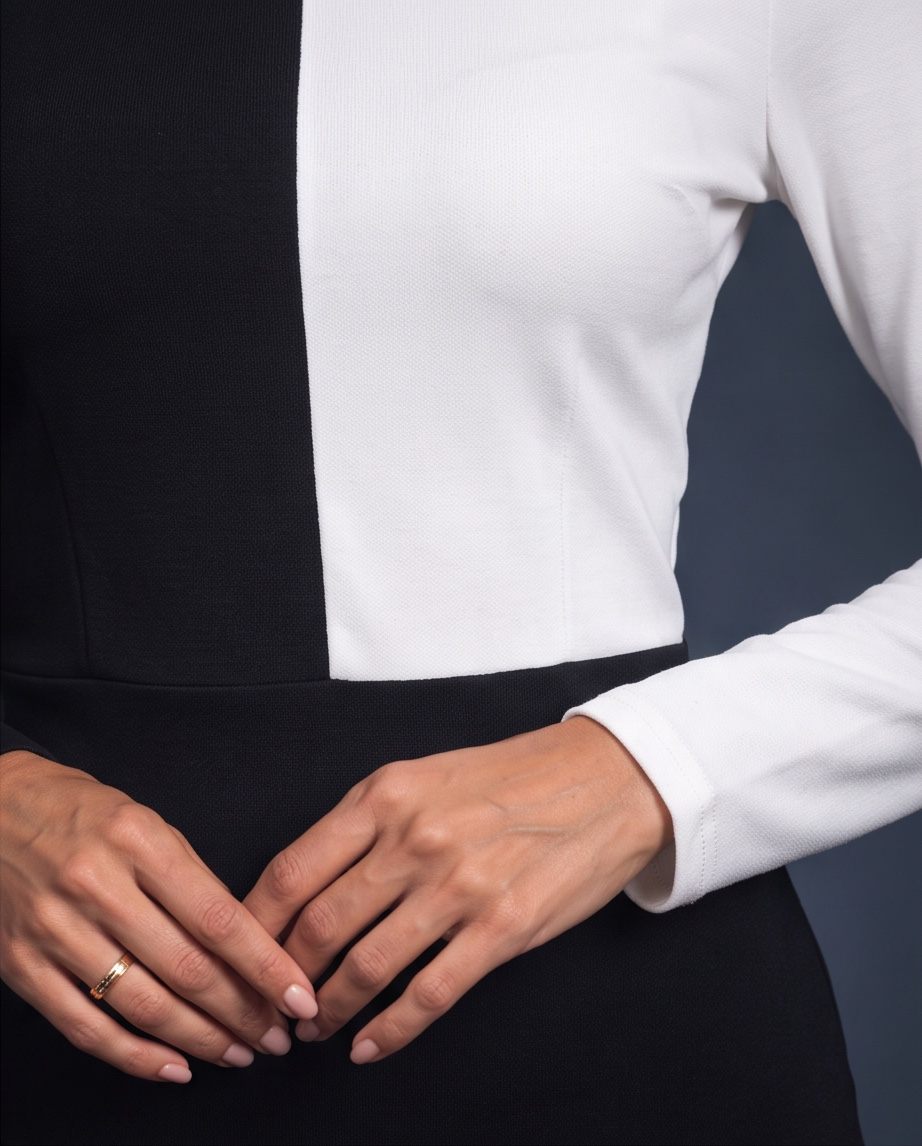 A close-up of a person wearing a color-block knit top with a vertical split of black and white, paired with a black skirt. The person's hands are clasped in front, and a gold ring is visible on one finger.