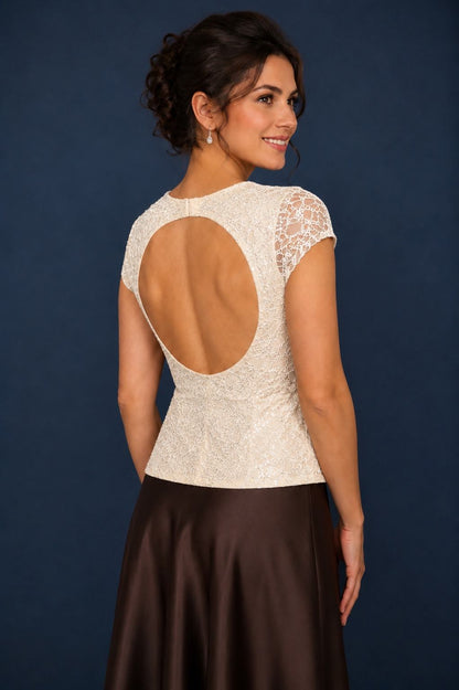 A woman shown from the back wearing a cream lace top with a large open back and cap sleeves, paired with a dark brown satin skirt, against a dark blue studio background.