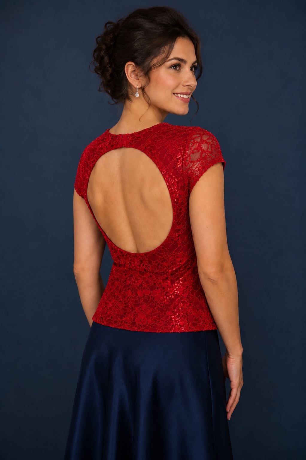 A woman shown from the back wearing a red lace dress with a large open back and short cap sleeves, paired with a navy satin skirt, against a dark blue studio background.