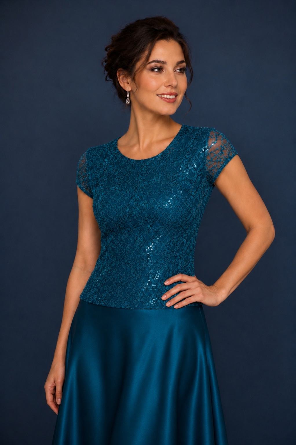 A woman modeling a teal blue lace top with short cap sleeves and a matching satin skirt against a dark blue studio background.
