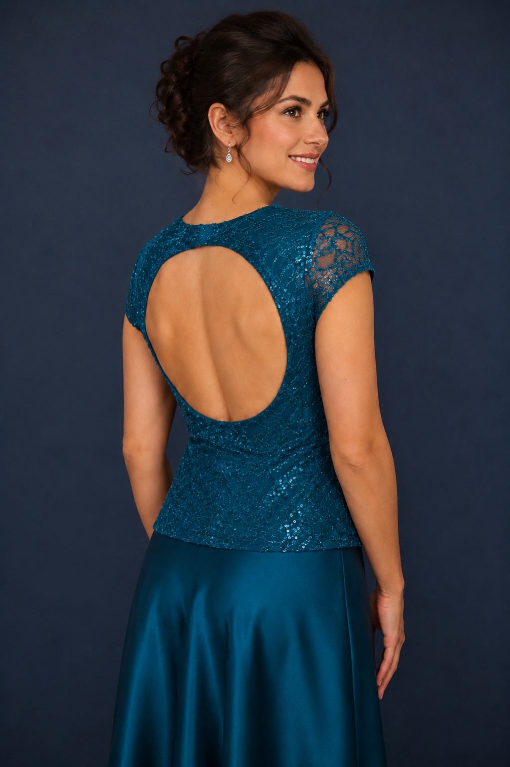 A woman shown from the back wearing a teal lace dress with a large open back and a satin skirt, posed against a dark blue studio background.