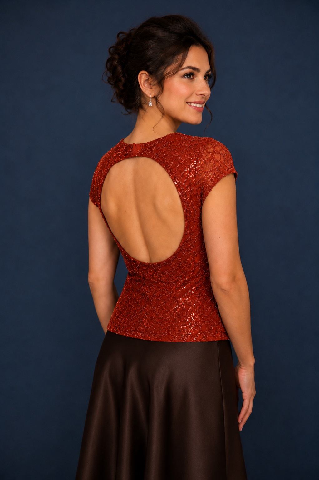A woman modeling a red sequined evening gown from the back, featuring a large open back and short cap sleeves, paired with a dark brown satin skirt against a dark blue background.