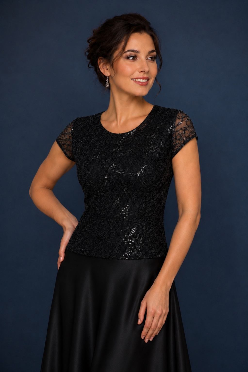 A woman modeling a black short-sleeve lace top with a sequined textured bodice, paired with a black satin skirt, set against a dark blue studio background.