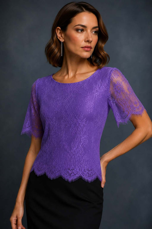A woman wearing a purple lace short-sleeve blouse with a scalloped hem and sheer lace sleeves, paired with a black skirt, posed against a dark gray studio background.