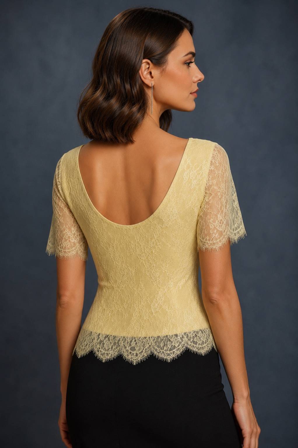 A woman shown from the back wearing a pale yellow lace short-sleeve top with a deep scoop neckline and a scalloped lace hem, paired with a black skirt against a dark blue-gray studio background.