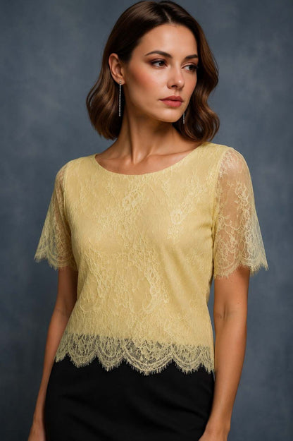 A woman wearing a pale yellow lace short-sleeve top with a scalloped hem and sheer lace sleeves, paired with a black skirt, against a dark gray studio background.