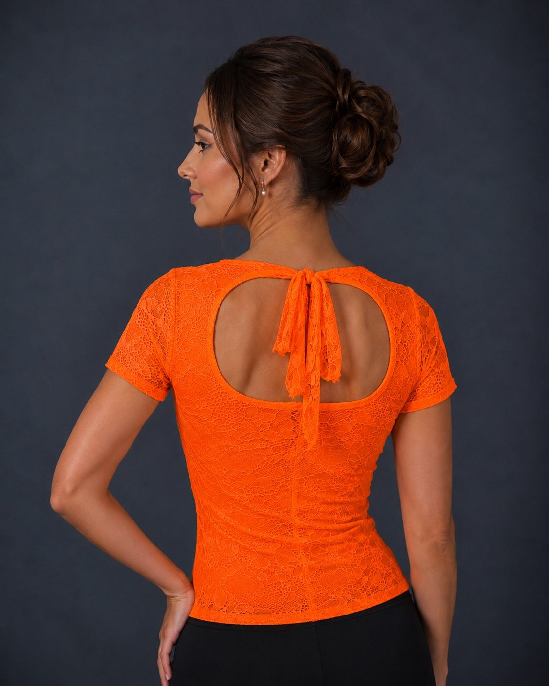 A woman shown from the back wearing a bright neon orange lace short-sleeve top with a large keyhole cutout and tie detail at the back, paired with black bottoms against a dark gray studio background.