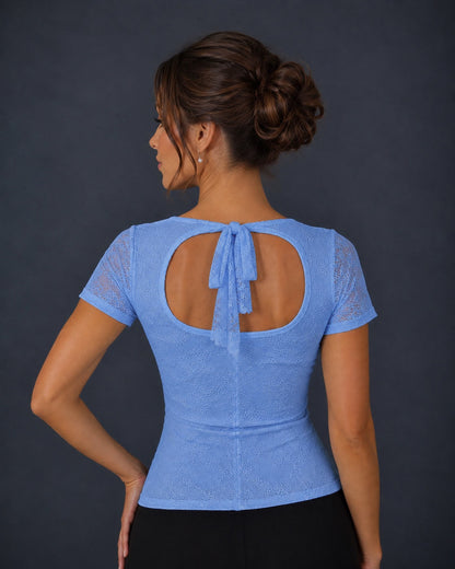 A woman shown from the back wearing a light blue short-sleeve lace top with a tie-back detail, paired with black bottoms, against a dark gray studio background.