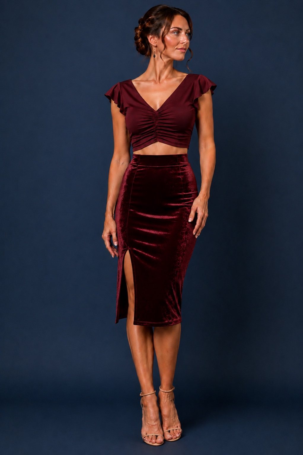 A woman models a two-piece velvet set consisting of a deep burgundy crop top with cap sleeves and a matching high-waisted midi skirt with a thigh-high slit, set against a dark blue studio backdrop.