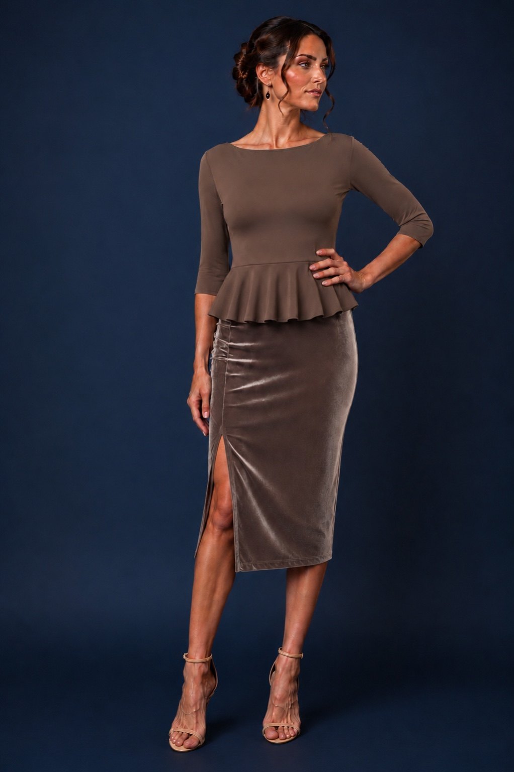 A woman models a taupe brown two-piece outfit consisting of a three-quarter sleeve peplum top and a matching velvet pencil skirt with a thigh-high slit, set against a dark blue studio background.