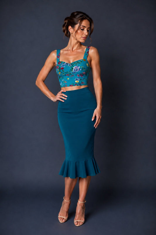 A woman models a two-piece outfit consisting of a teal blue floral crop top with floral embroidery and a matching teal blue flared midi skirt, paired with beige high-heeled sandals against a dark gray studio background.
