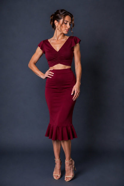 A woman models a two-piece burgundy set consisting of a ruched crop top with cap sleeves and a matching high-waisted flared skirt, posed against a dark gray studio backdrop.
