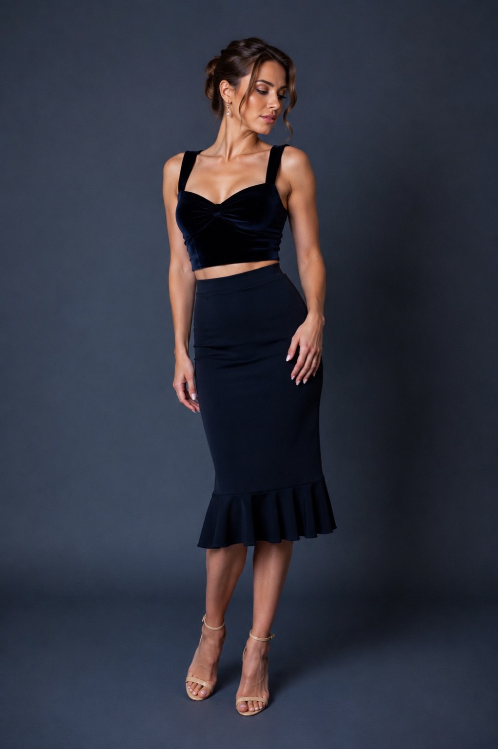 A woman modeling a two-piece black velvet and satin set consisting of a sweetheart crop top with wide straps and a high-waisted ruffled midi skirt, against a dark gray studio background.