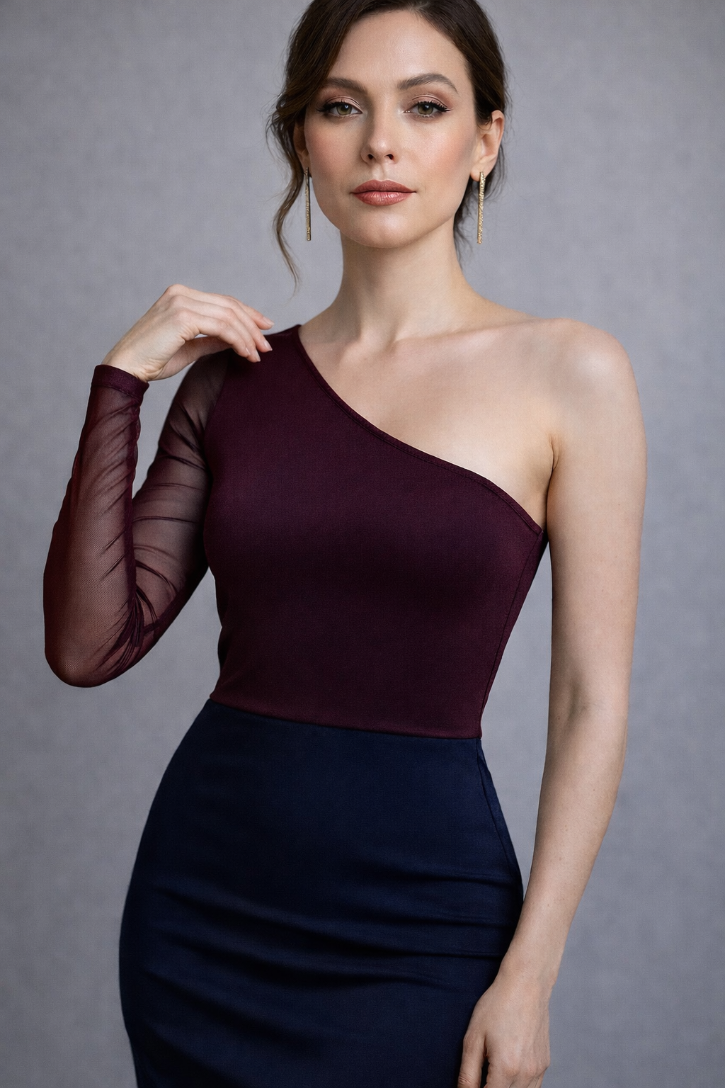 A woman modeling a one-shoulder dress with a deep burgundy mesh long sleeve and a dark navy blue fitted skirt, set against a neutral gray studio background.