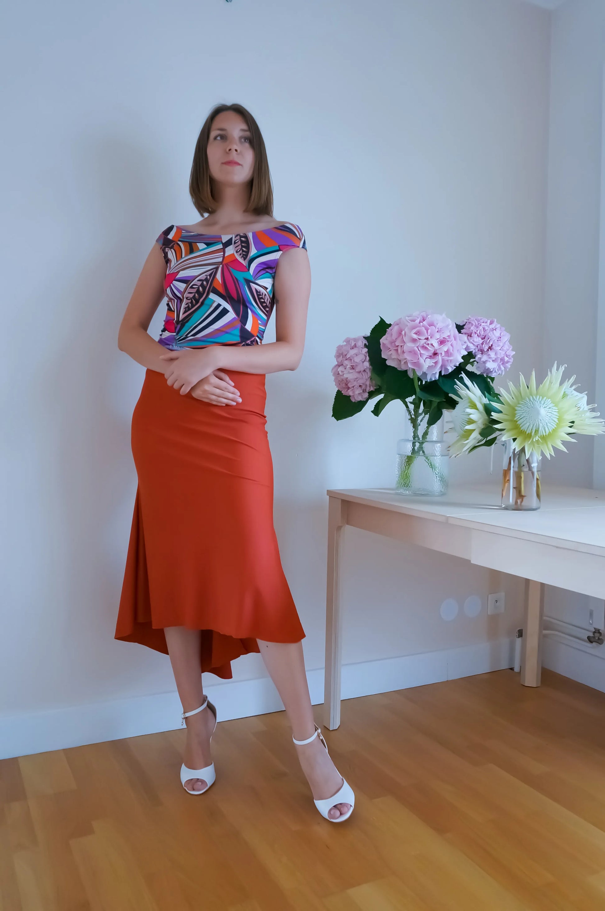Woman in a colorful tango top and orange tango skirt standing in a room with a table and flowers.