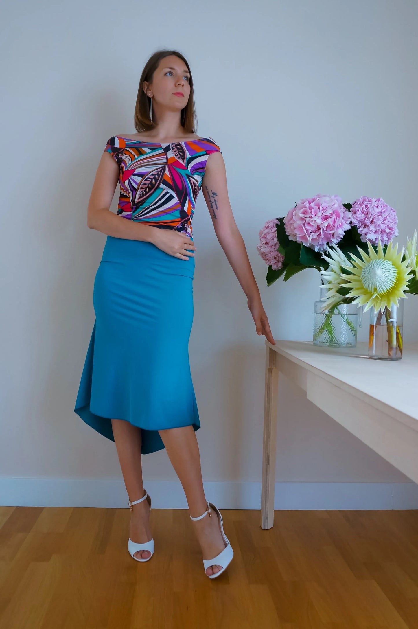 Woman in a colorful tango top and blue tango skirt standing next to a table with flowers against a white wall.