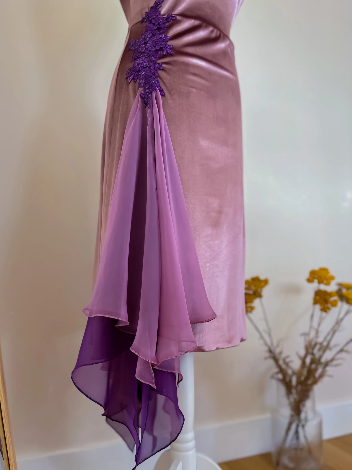 Purple evening tango gown with floral details on a white stand against a beige wall.